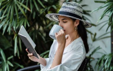Thoughtful Asian female in stylish white shirt and straw hat reading notes and drinking cup of coffee while sitting alone near green plants